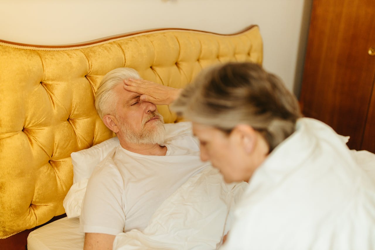 An elderly man in bed being checked for fever by a caregiver, suggesting illness or resting state.